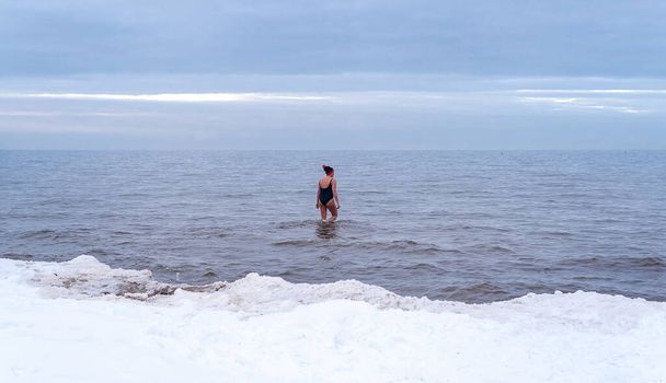 Μια γυναίκα με κόκκινο καπέλο κολυμπάει στη Βαλτική θάλασσα το χειμώνα καλυμμένη με χιόνι στο φόντο του πάγου. Υγιεινός τρόπος ζωής Το χειμερινό κολύμπι. Σπάνιο φαινόμενο. Λετονία, Jurmala - Φωτογραφία, εικόνα