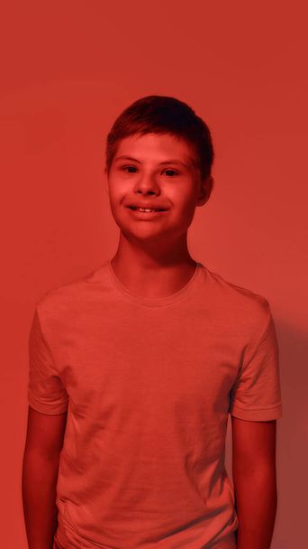 Portrait of joyful teenaged disabled boy with Down syndrome smiling at camera while posing isolated over red light background - Fotografie, Obrázek
