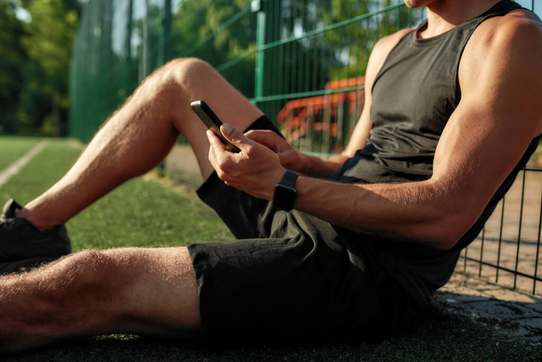 Обрезанный снимок молодого спортсмена в наушниках, сидящего на поле и слушающего музыку в солнечный летний день - Фото, изображение