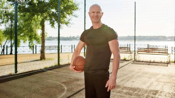 Happy adult basketball player smiling at camera while posing with a basketball on the court on a sunny day - Foto, immagini