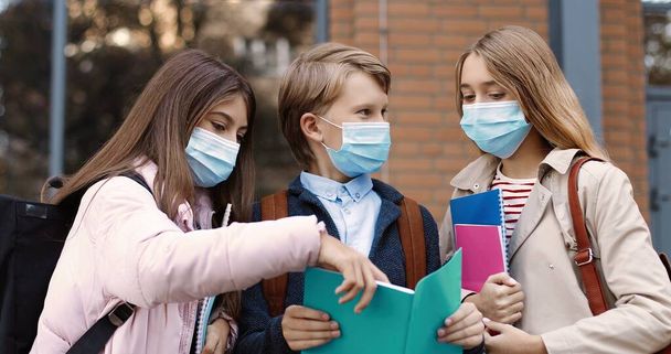 Portrait of joyful Caucasian school children standing outdoor and talking. Happy beautiful teen girls in masks and boy with copybook reading and chatting before lessons. Education concept - Foto, imagen