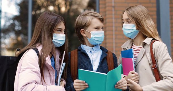Portrait of joyful Caucasian school children standing outdoor and talking. Happy beautiful teen girls in masks and boy with copybook reading and chatting before lessons. Education concept - Foto, Imagem