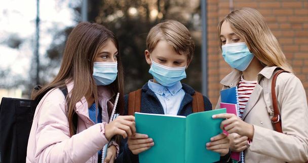 Portrait of joyful Caucasian school children standing outdoor and talking. Happy beautiful teen girls in masks and boy with copybook reading and chatting before lessons. Education concept - Foto, Imagen