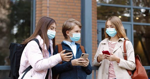 Portrait of Caucasian teen school kids standing near school and typing on smartphones. Beautiful girls in masks talking and showing boy something on cellphone. Classmates with backpacks School concept - Foto, Bild