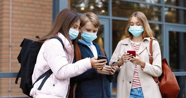 Portrait of Caucasian teen school kids standing near school and typing on smartphones. Beautiful girls in masks talking and showing boy something on cellphone. Classmates with backpacks School concept - Foto, Bild