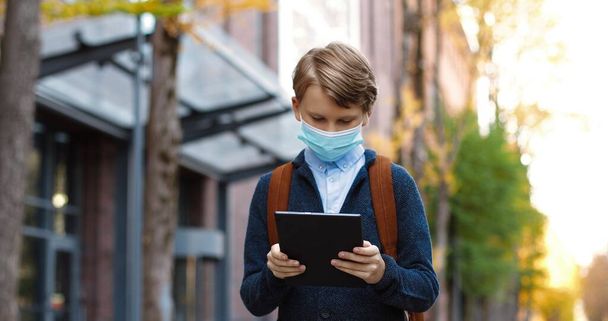 Portrait of cute teen boy in medical mask walking and tapping on tablet outdoors. Caucasian male pupil with backpack browsing and typing on device in city. School student. Knowledge concept - 写真・画像