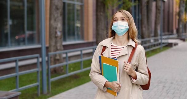 Portrait of cheerful young teen girl in mask walking outdoors on street with backpack and copybooks. Caucasian beautiful happy female school pupil after classes in quarantine. Education concept - Photo, image