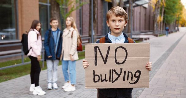 Portrait of cute teen schoolboy standing outdoors with "No bullying" sign while Caucasian kids laughing at him on background. Male junior student near school against mocking and abusing School concept - Foto, Imagem