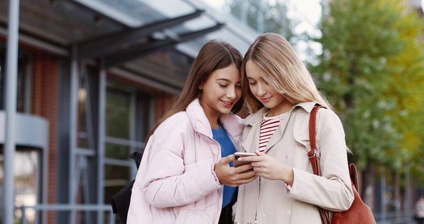 Portrait of Caucasian joyful beautiful school girls chatting while standing outdoors in good mood and typing on smartphone. Happy female pupils talking and using cellphone. Teenage concept - Foto, Imagem