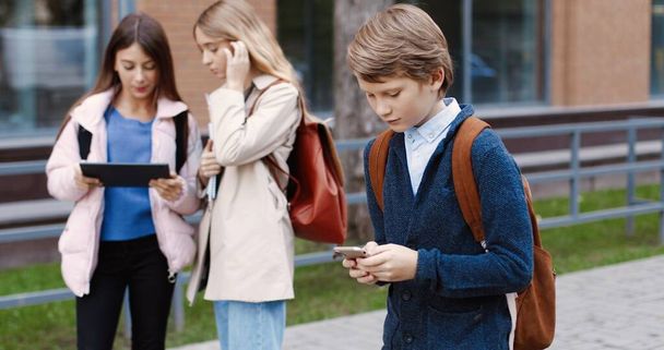 Portrait of Caucasian school student boy going to school and typing on cellphone outdoors. Male pupil texting and tapping on smartphone. Girls tapping on tablet on background. Pupils concept - Foto, Imagem