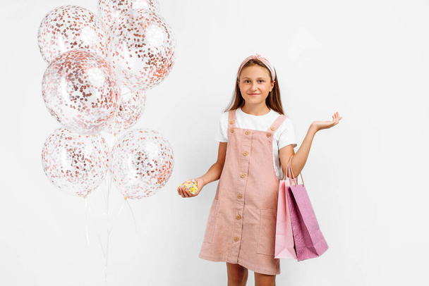 Happy stylish little girl in pink dress with balloons and with shopping bags looking at camera on isolated white background, shopping concept, - Фото, изображение