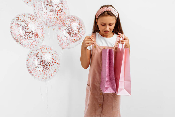 Счастливая привлекательная маленькая девочка держит сумки и воздушные шары изолированы на белом фоне - Фото, изображение