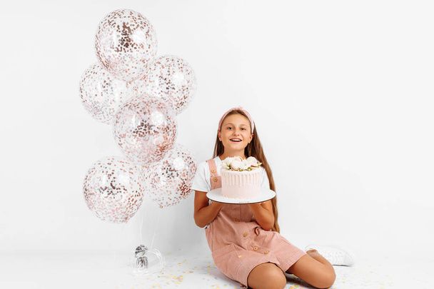 Cheerful beautiful child with balloons and big beautiful cake celebrates his birthday sitting on isolated white background - Photo, image
