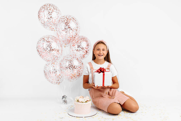 Happy young girl, with balloons, big beautiful cake and gift, sitting on isolated white background - Fotografie, Obrázek