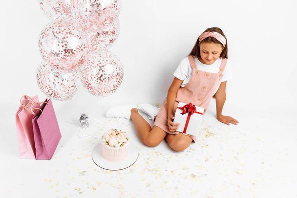 Happy young girl, with balloons, big beautiful cake and gift, sitting on isolated white background - Fotó, kép