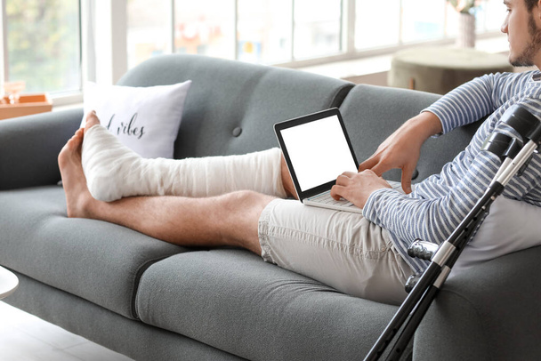 Young man with broken leg using laptop at home - Zdjęcie, obraz