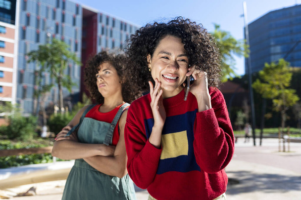 Young female is bored standing in the street while her woman friend talking on the phone and smiling - Foto, Imagem