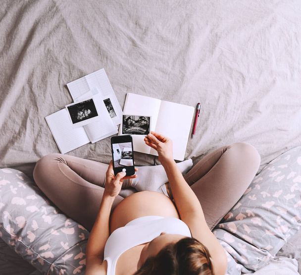 Pregnant woman holding in hands smartphone with empty white display. Close-up pregnant belly of expectant mother with ultrasound photos and medical test reports. Concept of pregnancy, gynecology. - Photo, image