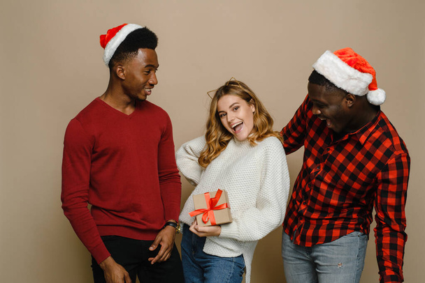 Twee zwarte mannen en een blanke vrouw vieren Kerstmis en Nieuwjaar op een beige achtergrond en kijken naar een geschenk. Indoor portret van beste vrienden klaar voor vakantie feest met helder cadeau - Foto, afbeelding
