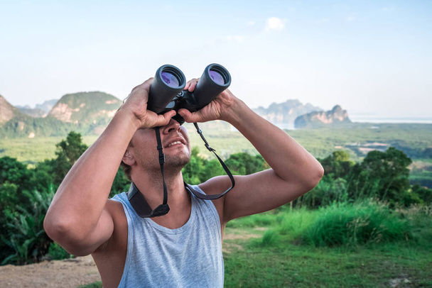 A young man looks into the sky through binoculars. A traveler on the background of wild nature. - Фото, изображение