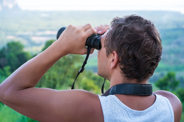 A young man looks through binoculars from the top on the background of the forest - Фото, изображение