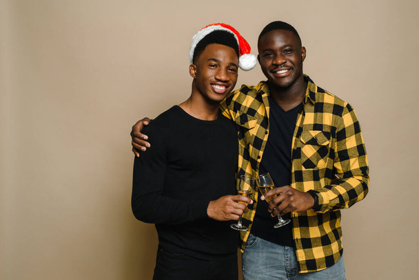 Glückliches Familienporträt zu Weihnachten, schwules Männerpaar auf beigem Hintergrund. Ein Paar verliebter schwarzer Männer klirrt mit Champagnergläsern. Prost. Feiern Sie Weihnachten und Neujahr - Foto, Bild