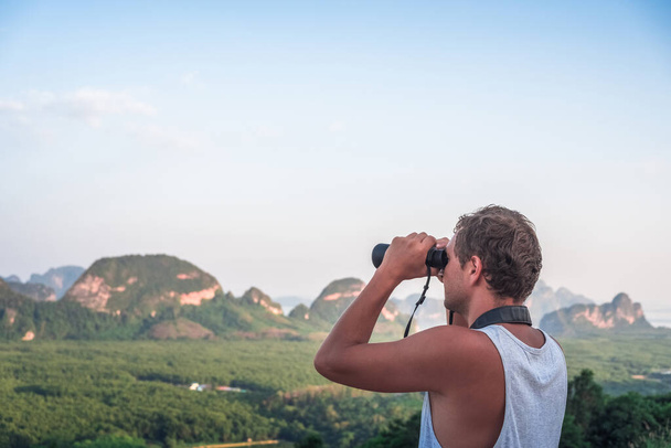 A young man in a white T-shirt looks from the top into the distance through binoculars against the backdrop of wildlife, sky, mountains and sea - Foto, imagen
