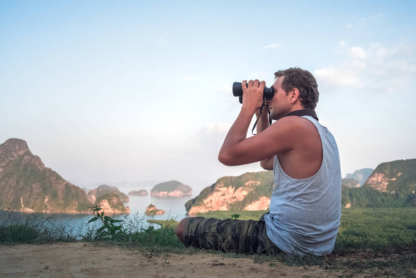 A young man in a white T-shirt sits on the top and looks into the distance through binoculars at a beautiful seascape and rocks - Photo, image