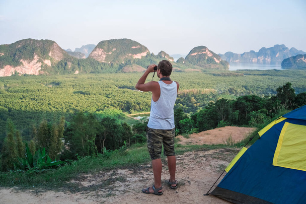 A young man in a white T-shirt stands on the top next to the tent and looks into the distance through binoculars, at the wildlife - Fotoğraf, Görsel