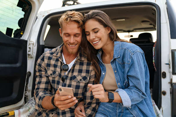 Beautiful couple sitting at the back of car and using smart phones - Фото, изображение