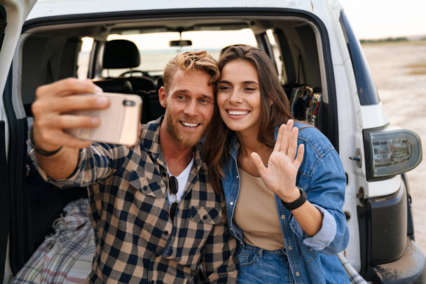 Young couple in love making selfie using smart phone while sitting on a back of car - Фото, зображення