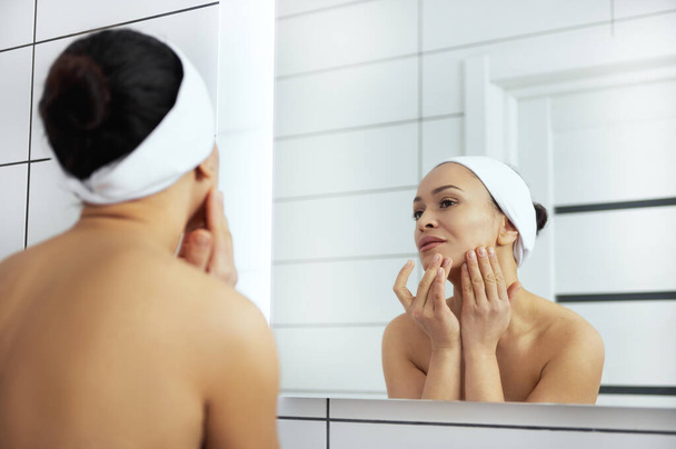 Die junge Frau berührt ihr Gesicht beim Betrachten der Spiegelung im Spiegel - Foto, Bild