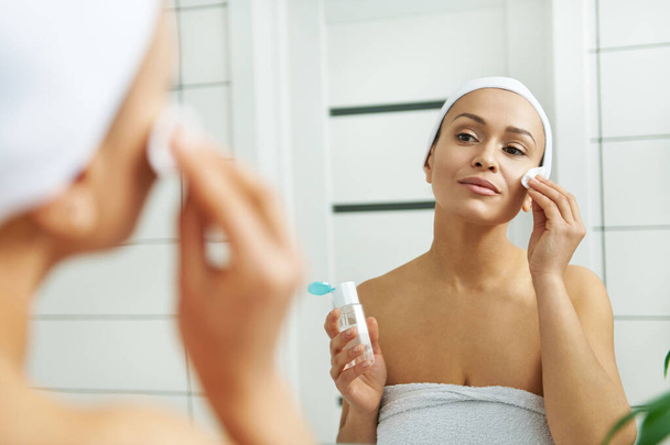De vrouw veegt gezicht met lotion met behulp van een wattenschijfje in de voorkant van een spiegel - Foto, afbeelding