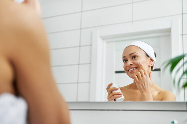 Женщина, использующая средство по уходу за кожей лица, глядя на свое отражение в зеркале ванной комнаты - Фото, изображение