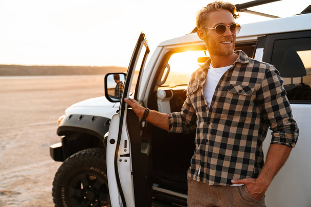 Handsome confident young man posing while standing at his car door at the beach - Photo, image