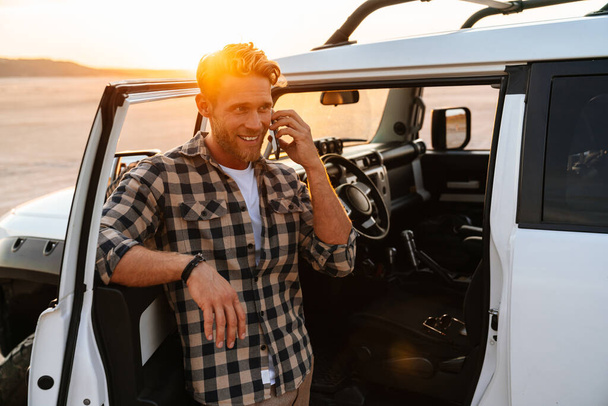 Handsome young man using mobile phone while standing at the car door at the beach, talking - 写真・画像