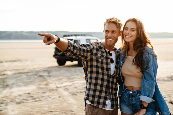 Young happy loving couple outdoors at beach near car walking, embracing, pointing away - Photo, image