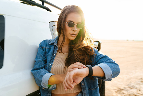 Attractive young woman leaning on a car while standing at the beach, looking at smart watch - Fotoğraf, Görsel