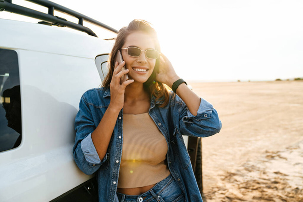 Attractive young woman leaning on a car while standing at the beach, using mobile phone, talking - Foto, imagen