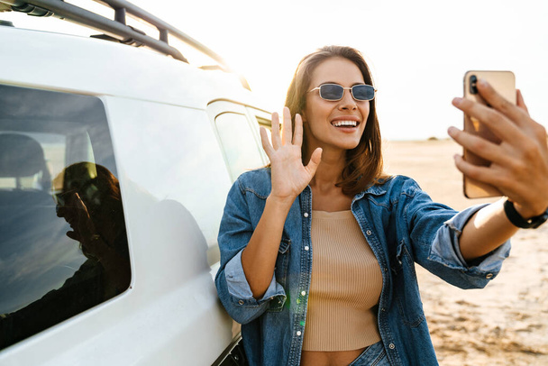 Attractive young woman leaning on a car while standing at the beach, using mobile phone, taking a selfie - Foto, afbeelding