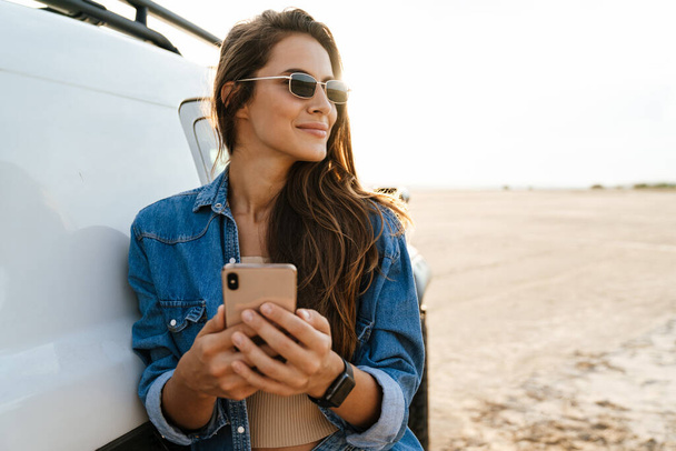 Attractive young woman leaning on a car while standing at the beach, using mobile phone - Φωτογραφία, εικόνα