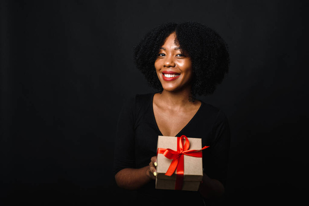 Glückliche schwarze junge Frau mit Afro hält ein Weihnachtsgeschenk in den Händen und lächelt. Bescherung und Weihnachtseinkauf. Schwarzer Freitag - Foto, Bild