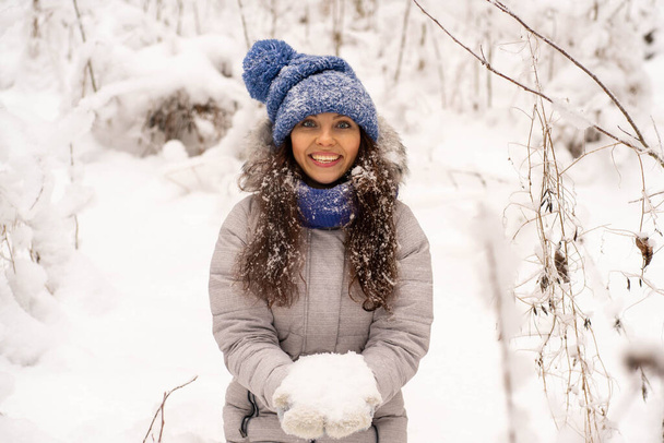 Beautiful happy laughing young woman in a winter hat with gloves and a scarf covered with snow flakes. Winter forest landscape background. - Foto, Imagem