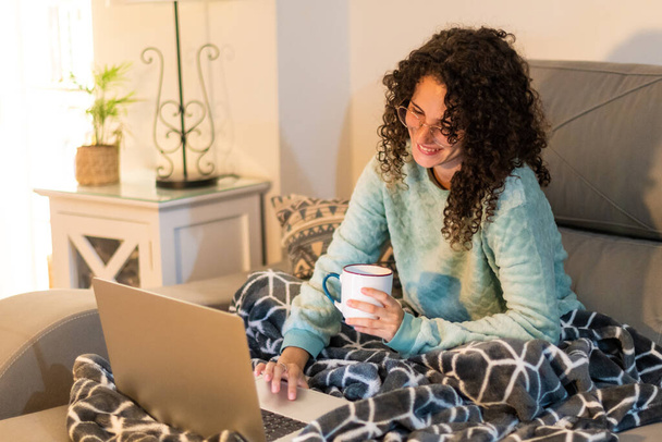 Archivbild einer jungen Frau mit Brille und lockigem Haar zu Hause, die es sich mit Laptop auf der Couch gemütlich macht. Kaukasische Frau trinkt einen Kaffee mit Winterschlafanzug und weicher Decke. - Foto, Bild