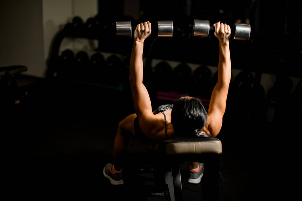 athletic brunette woman training with dumbbells lying on bench in gym. - Photo, Image