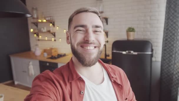 Handheld shot van vrolijke blanke man met volle baard houden mobiele apparaat in uitgestrekte hand en lopen rond keuken thuis terwijl video chatten met iemand gelukkig, glimlachen en lachen - Video