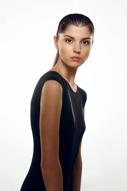 Vue de face d'un modèle de brune mince en robe noire élégante, aux cheveux serrés, posant sur un fond blanc. - Photo, image