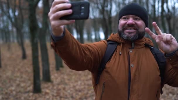Herbstlicher Mann mit schwarzem Hut und brauner Jacke im Wald. Macht ein Selfie am Telefon. Konzept der Form eines Bloggers in der Natur - Filmmaterial, Video