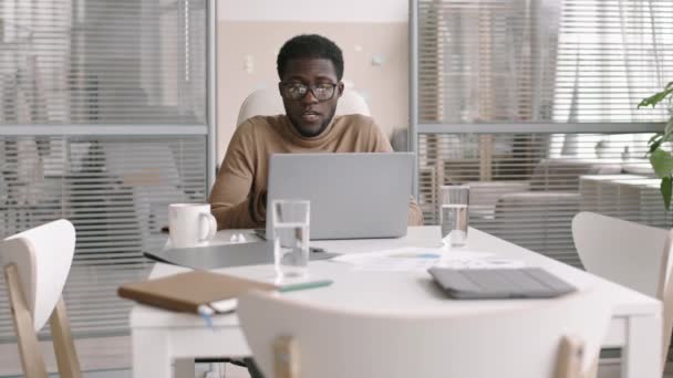 Zoom in op jonge knappe Afro-Amerikaanse zakenman die achter zijn bureau zit voor een laptop op kantoor en via een videoverbinding met zijn zakenpartner praat - Video