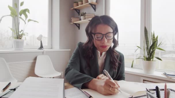 Waist-up POV shot of glamorous mixed race business lady in glasses and jacket sitting at desk in office, looking at camera, talking to client, listening, nodding and making notes - Footage, Video